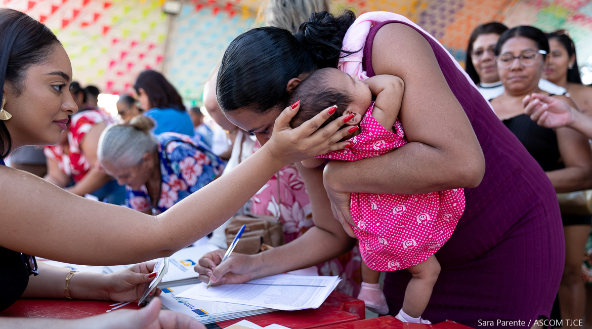 Funcionária segura a cabeça da criança para auxiliar a mãe, que assina o documento Funcionária segura a cabeça da criança para auxiliar a mãe, que assina o documento