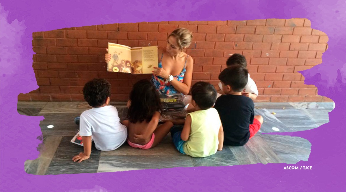 Foto de uma mulher sentada monstrando um livro de leitura para cinco crianças também sentadas Foto de uma mulher sentada monstrando um livro de leitura para cinco crianças também sentadas