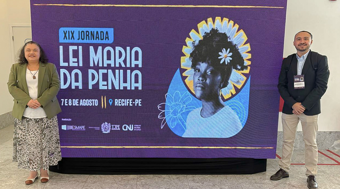 Foto com uma mulher e um homem e, entre eles dois, um cartaz da Lei Maria da Penha Foto com uma mulher e um homem e, entre eles dois, um cartaz da Lei Maria da Penha