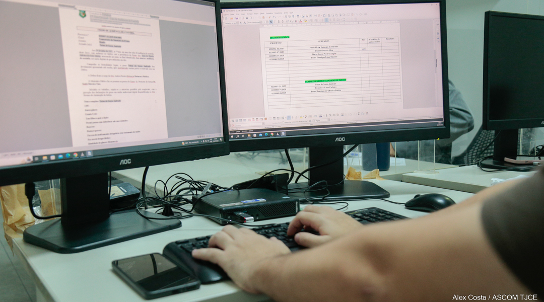 Mãos digitando em um teclado de computador diante de dois monitores