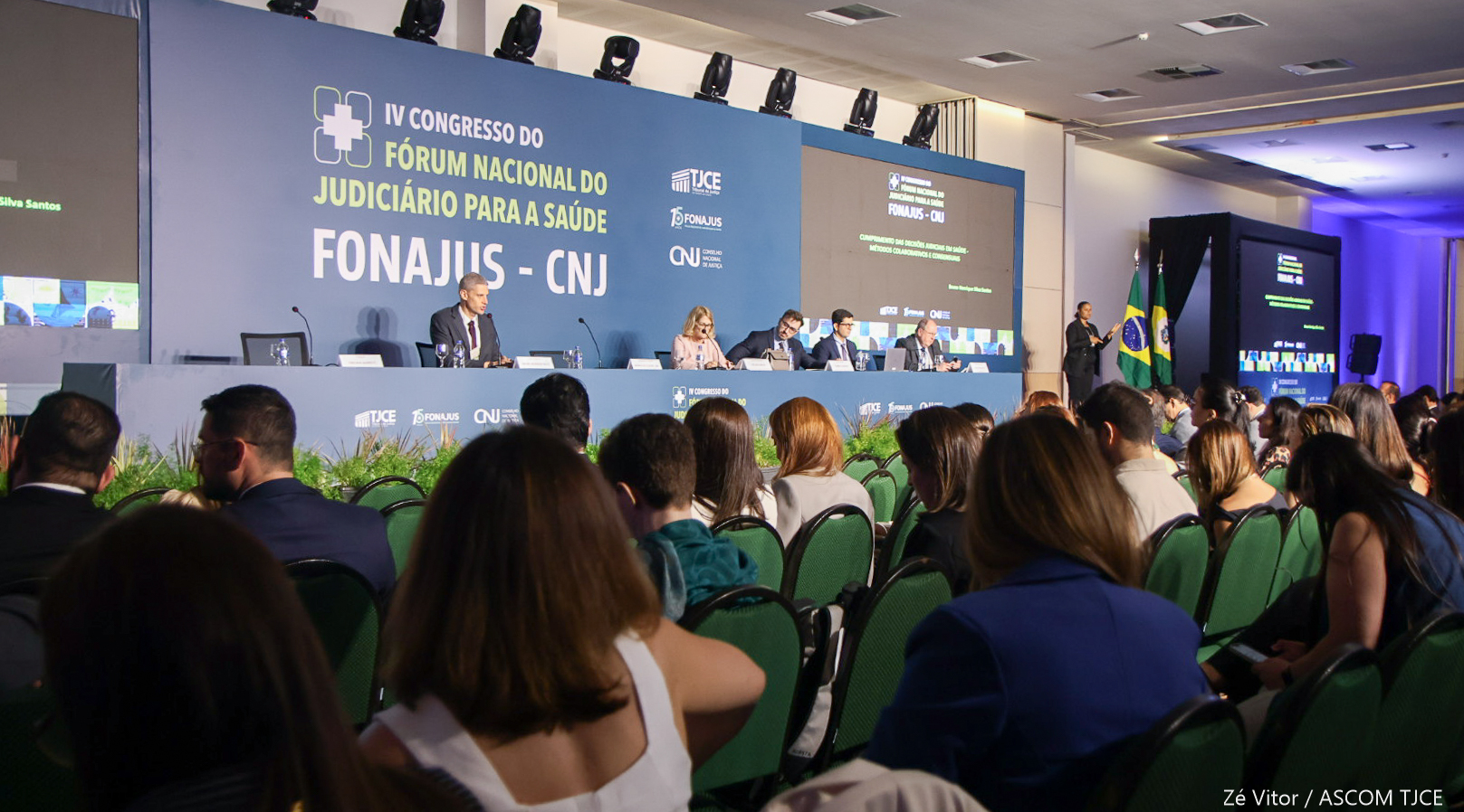 Palestrantes em mesa de debates de frente da plateia, que aparece de costas Palestrantes em mesa de debates de frente da plateia, que aparece de costas