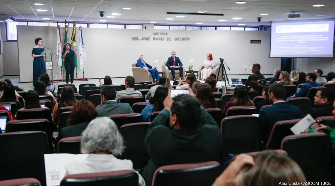 Foto no auditório com várias pessoas sentadas e uma mulher falando ao microfone