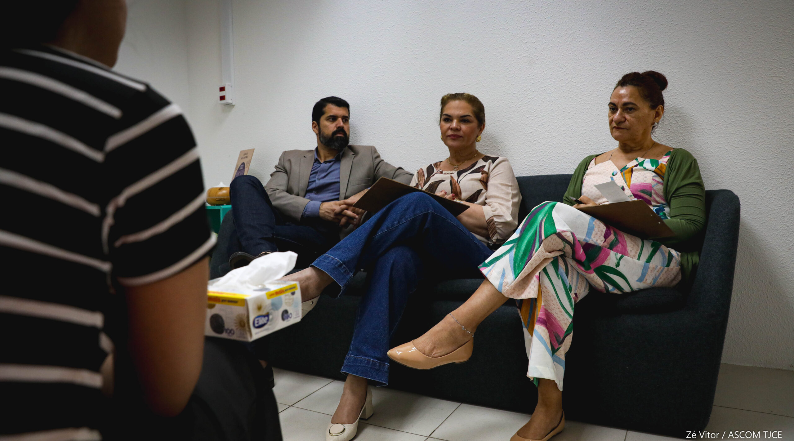 Um homem e duas mulheres, sentados em um sofá, de frente a uma pessoa de costas, também sentada Um homem e duas mulheres, sentados em um sofá, de frente a uma pessoa de costas, também sentada