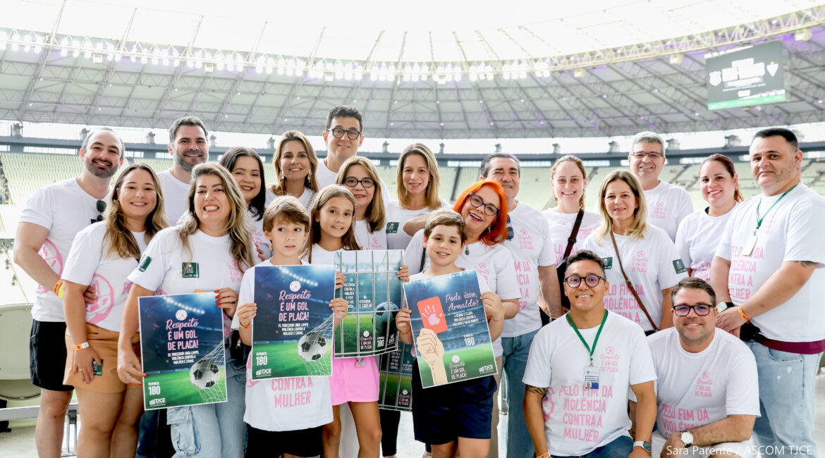 Adultos e crianças posam sorridentes com cartazes e camisetas brancas da campanha, próximos ao campo do estádio