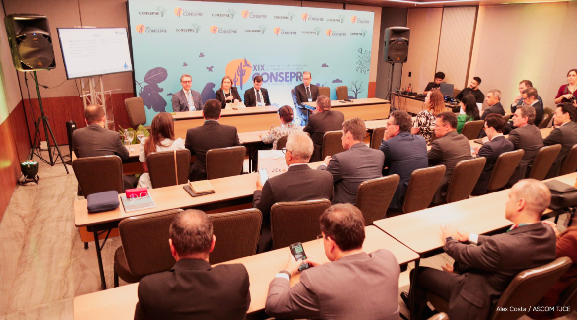 Foto com várias pessoas sentadas em um auditório e, ao fundo, um grande banner escrito Consepre
