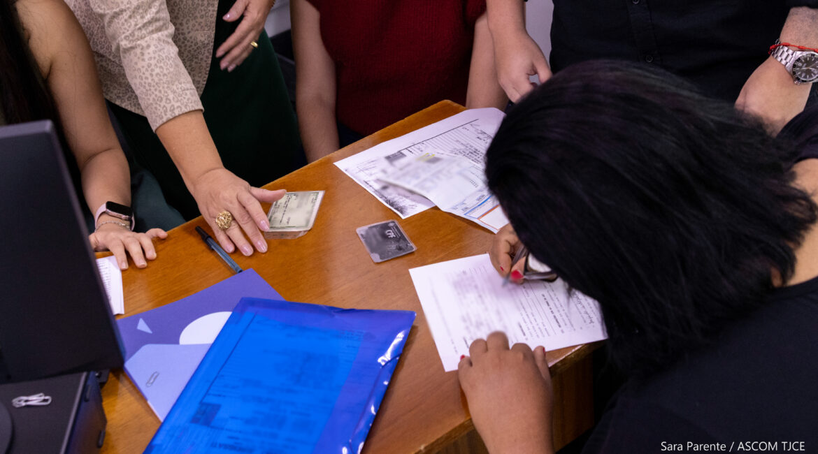 Foto de uma mesa mostrando mãos segurando documentos e umas pessoa, de costas, assinando um papel