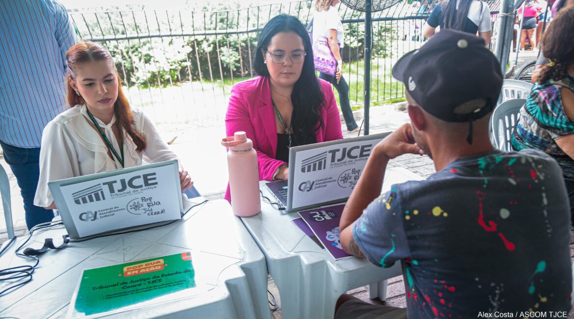 Foto de duas mulheres, sentadas, fazendo atendimento de um homem com boné na cabeça, de costas
