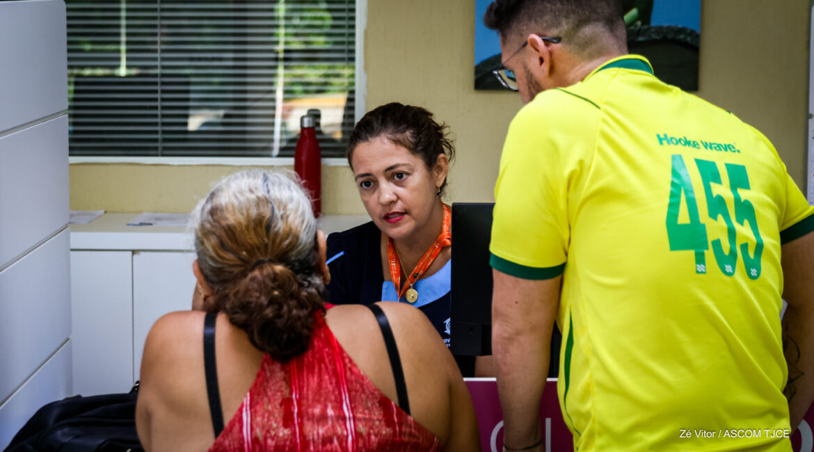 Foto com uma mulher sentada, fazendo o atendimento de duas pessoas
