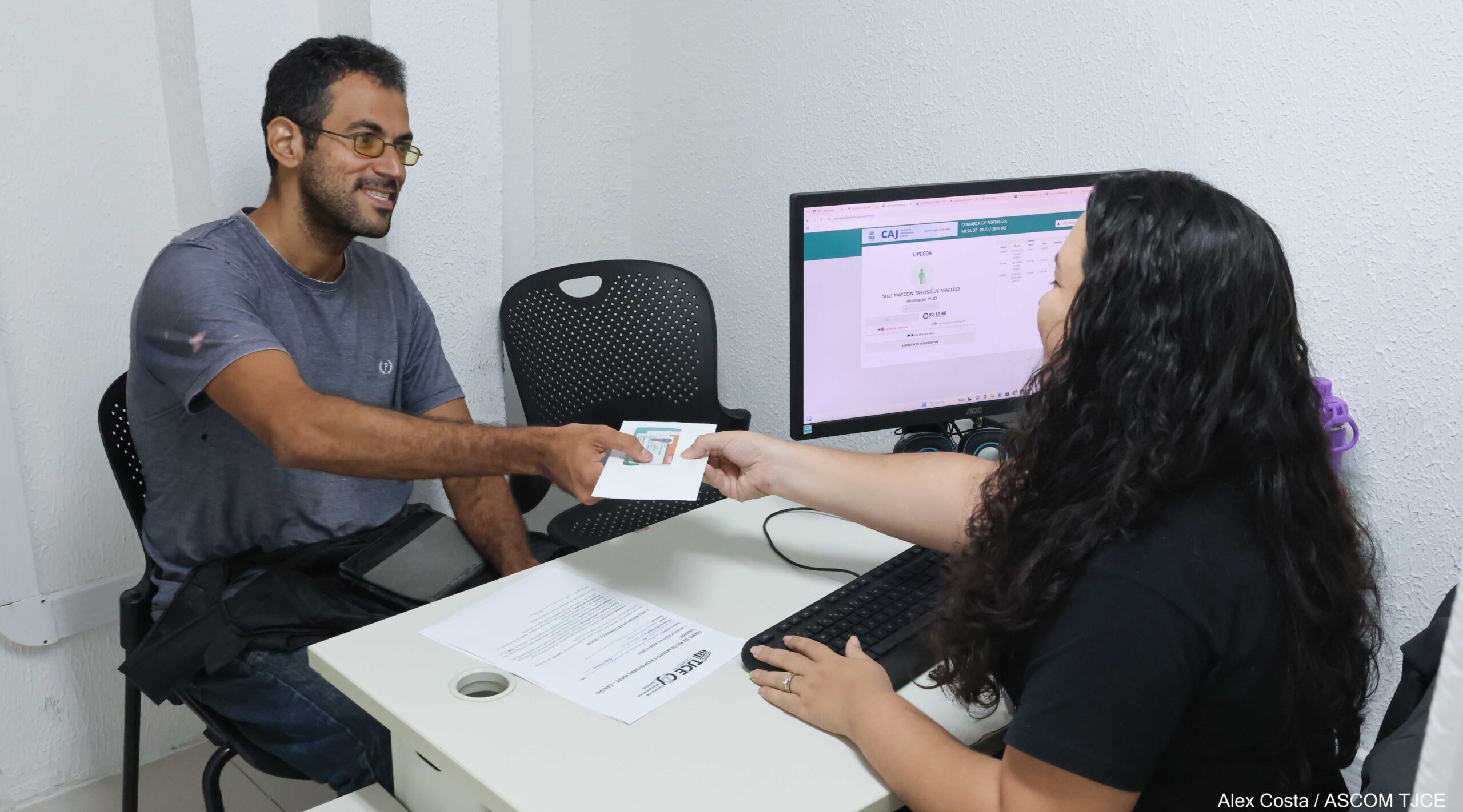 Foto com uma funcionária atendendo um homem numa sala. Ambos estão sentados e, no meio deles, aparece a tela de um computador