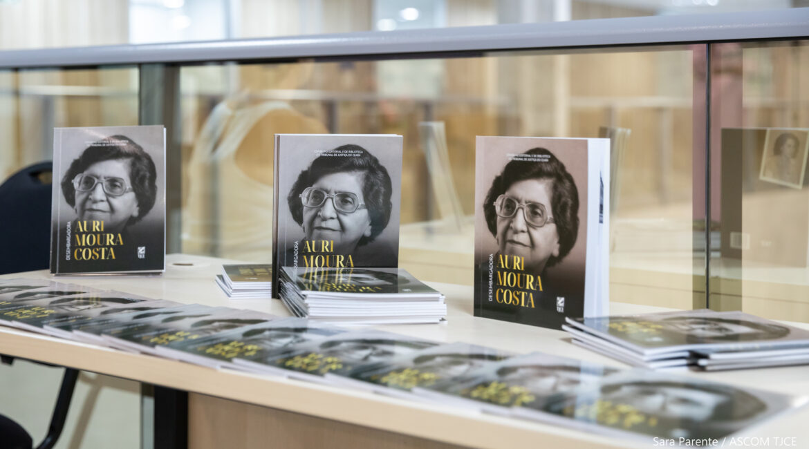 Foto da exposição de três livros de Auri Moura Costa, na vertical, e de vários na posição horizontal Foto da exposição de três livros de Auri Moura Costa, na vertical, e de vários na posição horizontal