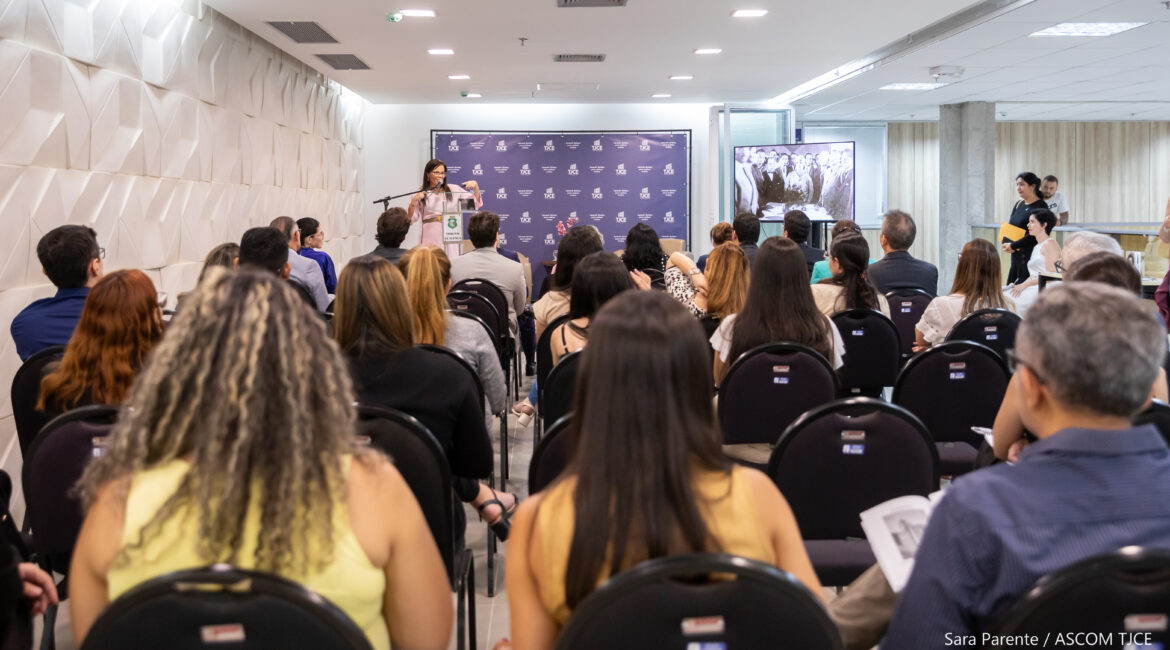 Foto com várias mulheres e homens sentadas(os) e, ao fundo, uma mulher em pé, falando ao microfone Foto com várias mulheres e homens sentadas(os) e, ao fundo, uma mulher em pé, falando ao microfone