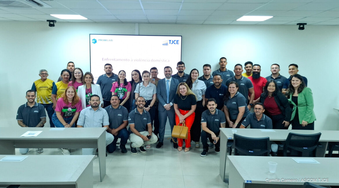 Foto com várias pessoas posando para foto, sorrindo, em uma sala ampla. Ao fundo, um painel grande, branco, com a logo do TJCE