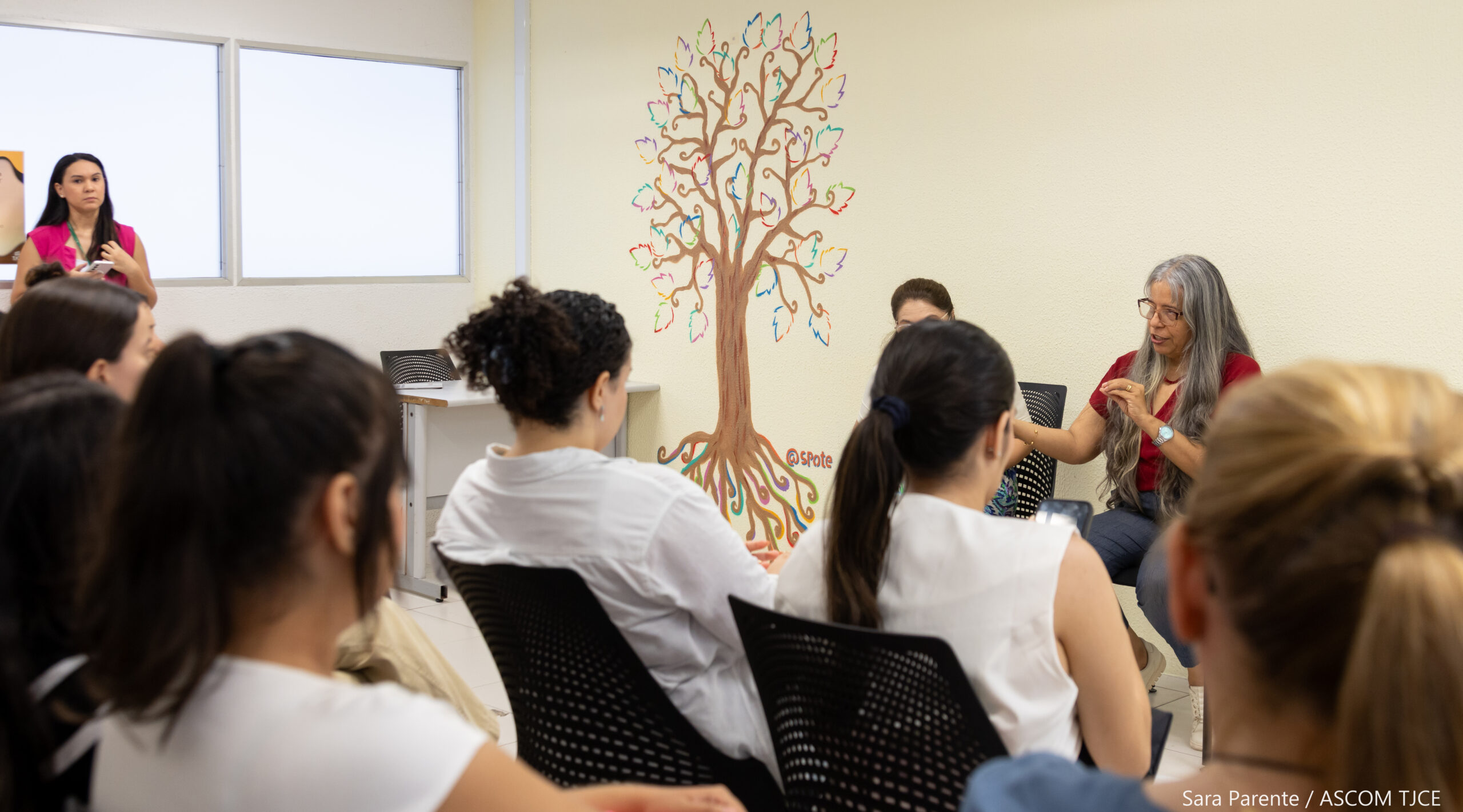 Foto com várias pessoas sentadas em uma sala e, ao fundo, uma mulher fazendo as explicações