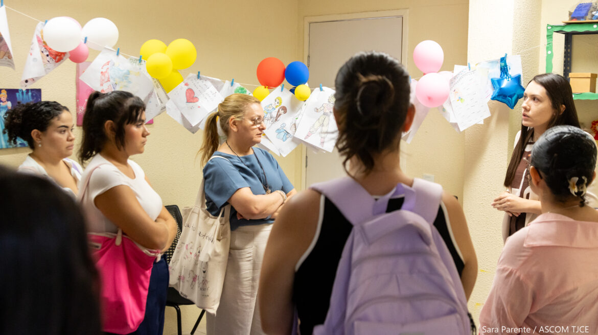 Foto com mulheres em uma sala, onde há também bolões de aniversários coloridos e papeis com dinâmicas pendurados em um fio Foto com mulheres em uma sala, onde há também bolões de aniversários coloridos e papeis com dinâmicas pendurados em um fio