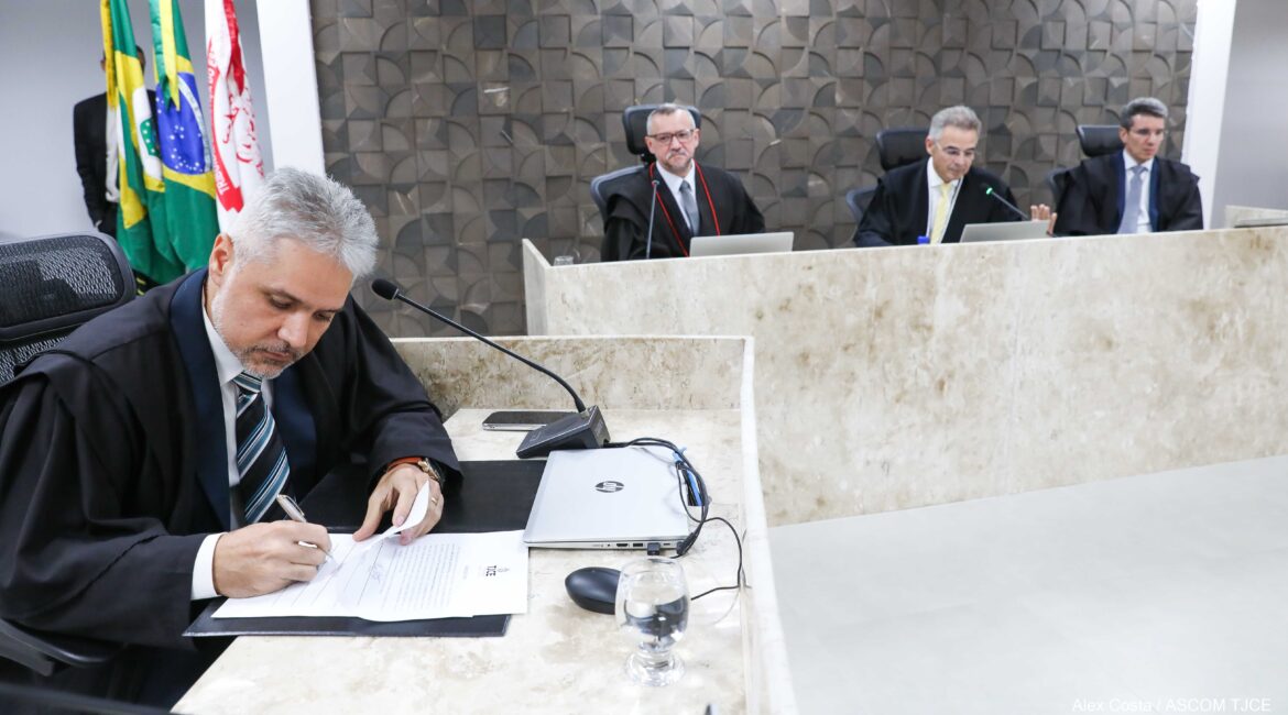 Foto de um homem, vestido com a toga, assinando um papel e, ao fundo, mais três homens sentados em uma bancada