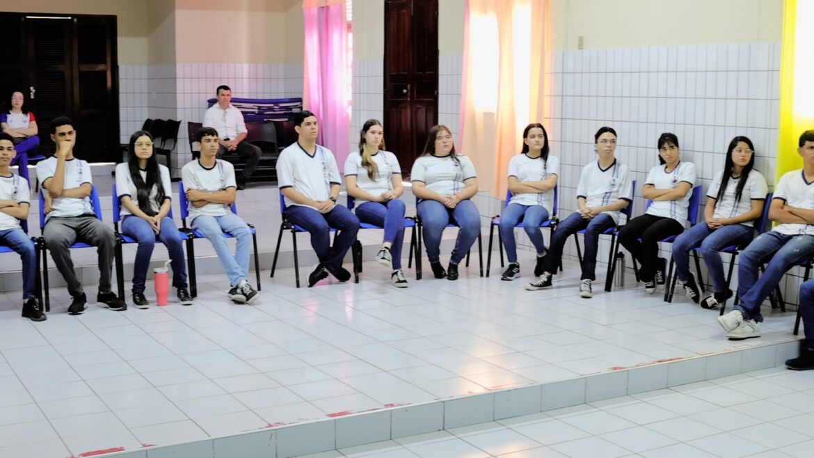 Foto com vários jovens sentados numa sala de aula no formato de um círculo. Eles estão vestidos com a farda do colégio, calça jeans azul e blusa branca