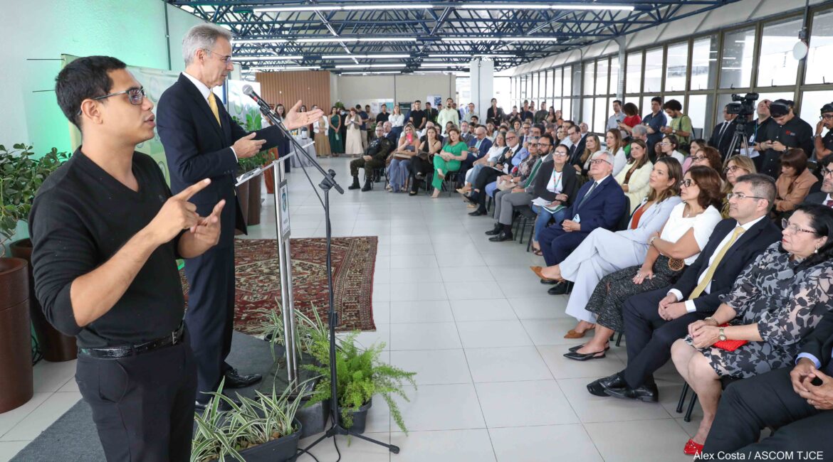 Foto de um homem falando em pé ao microfone para um público formado por homens e mulheres sentadas(os) Foto de um homem falando em pé ao microfone para um público formado por homens e mulheres sentadas(os)