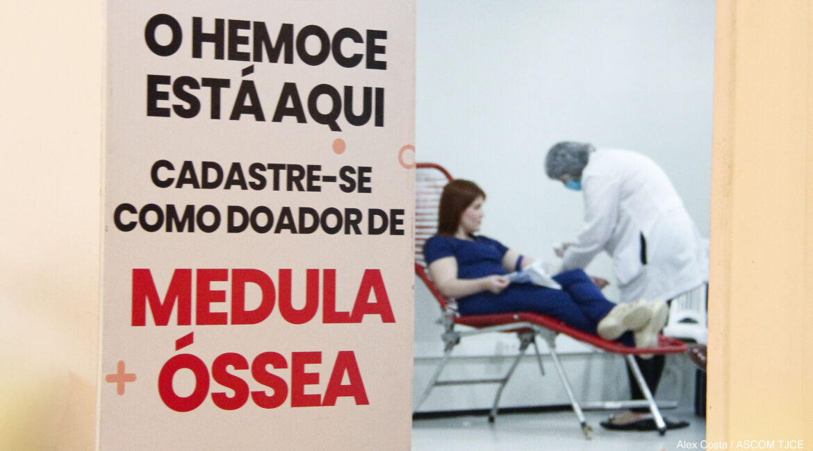 Foto de uma propaganda do Hemoce, disponibilizando cadastro para doação de medula óssea. Ao fundo, uma mulher sentada e uma profisisonal colhendo o sangue dela