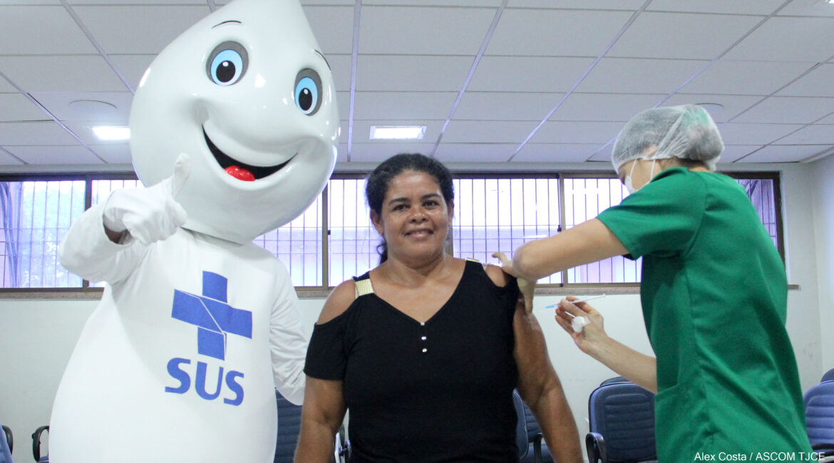 Foto de uma mulher sendo vacinada por outra mulher. Ao lado delas, um boneco inflável na cor branca, com o símbolo do SUS 