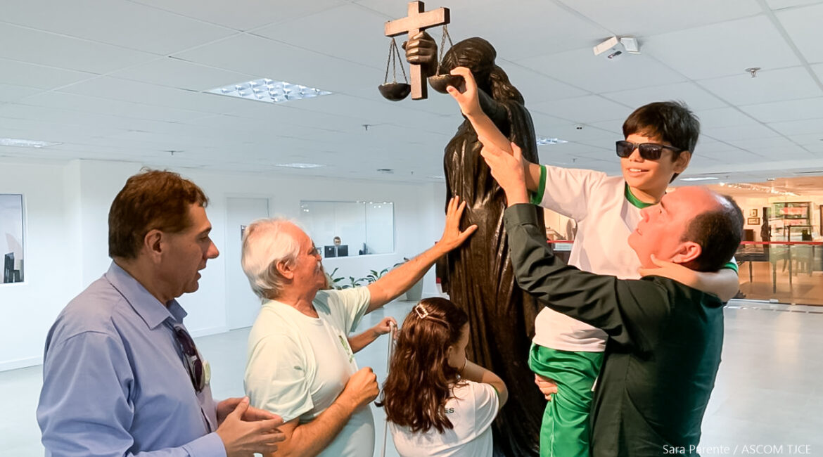 Foto de um homem que levanta nos braços uma criança para pegr na estátua da themys no hall do Tribunal. Ao lado dele, dois homens cegos e uma criança cega também tocam na estátua