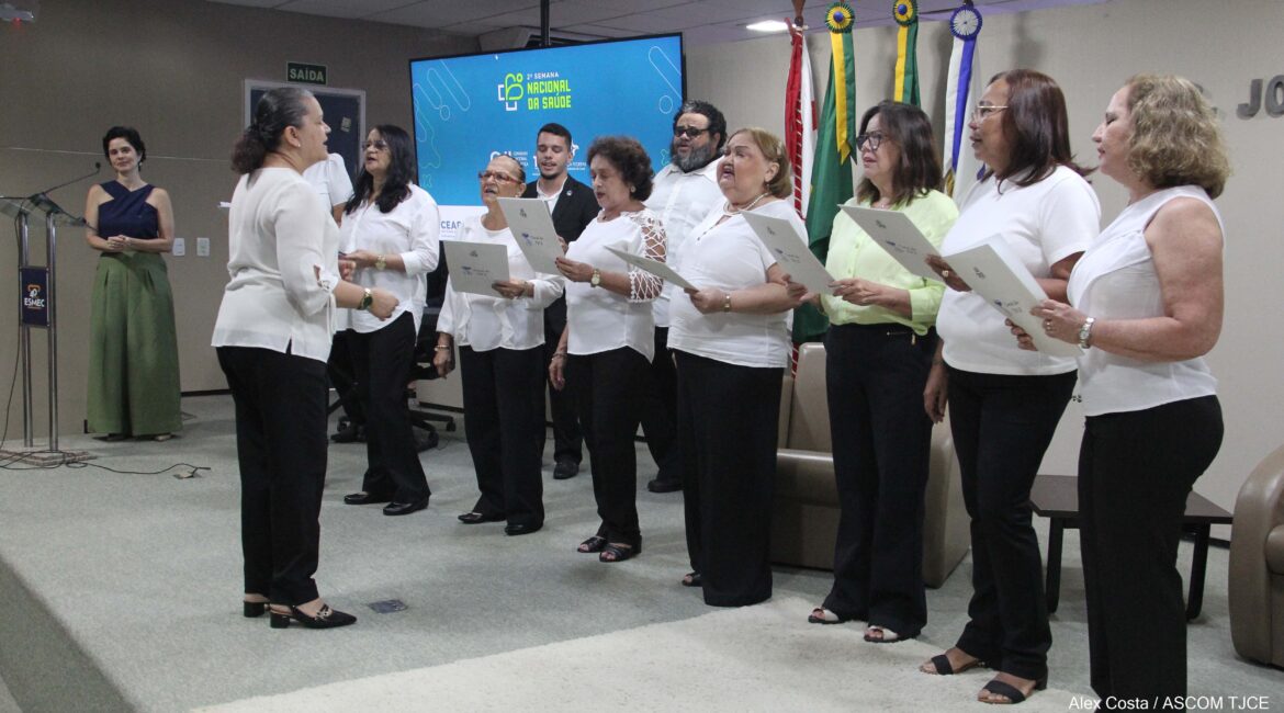 Foto com várias muheres e homens em cima de um palco, cantando, todos de branco Foto com várias muheres e homens em cima de um palco, cantando, todos de branco