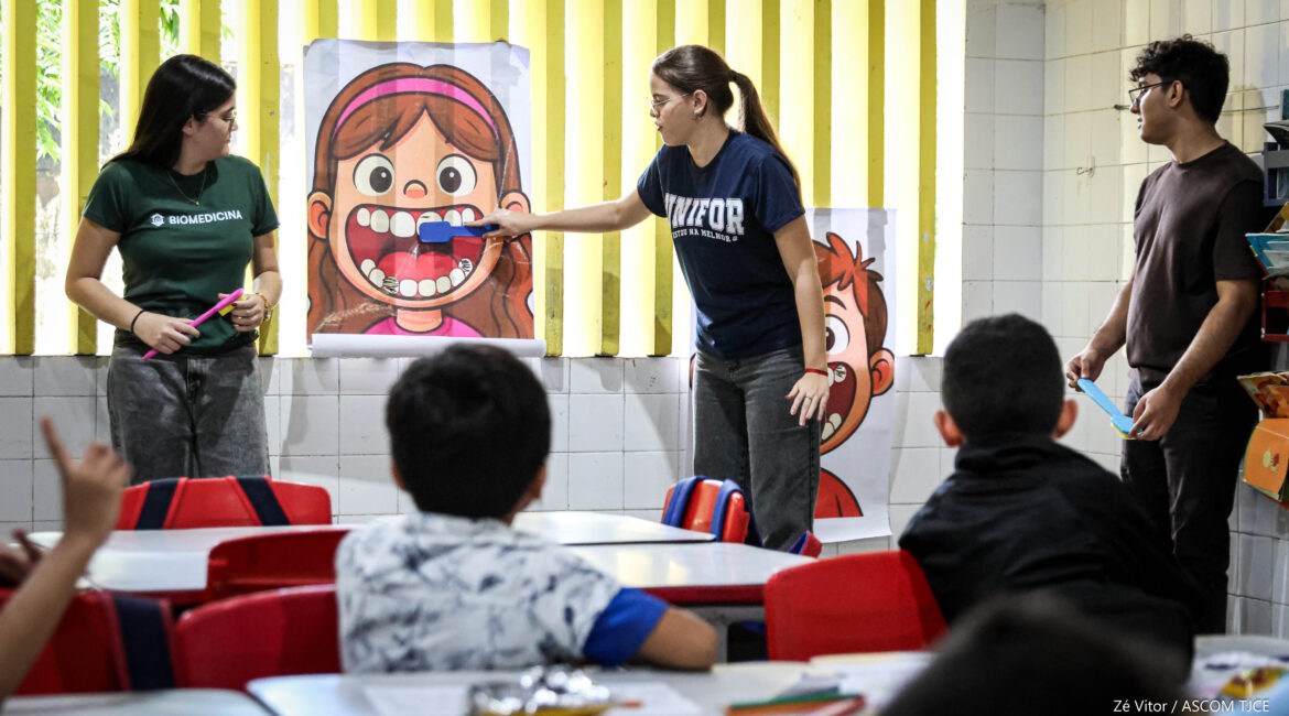 Foto de uma mulher simulando a escovação de dentes, em uma imagem de criança desenhada no papel. Ao lado dela, uma jovem e um jovem observando e, à frente dela, crianças sentadas, atentas, ouvindo as orientações