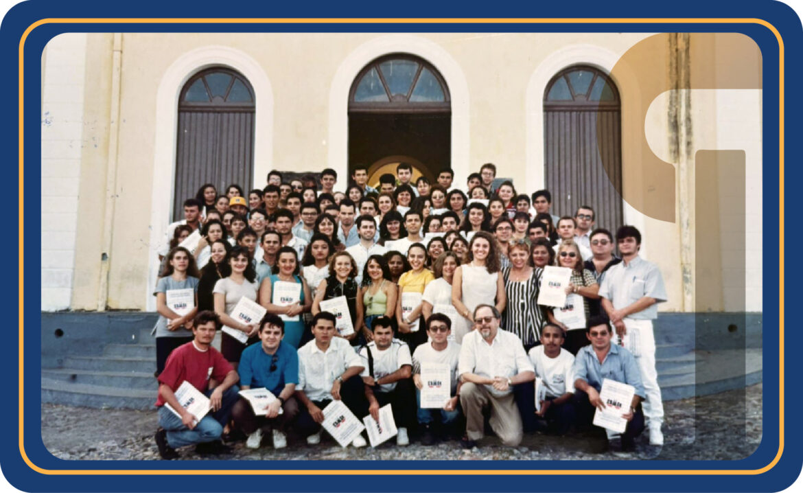Imagem com vários homens e mulheres posando para foto, sorrindo, e ao fundo, a fachada de um casarão Imagem com vários homens e mulheres posando para foto, sorrindo, e ao fundo, a fachada de um casarão