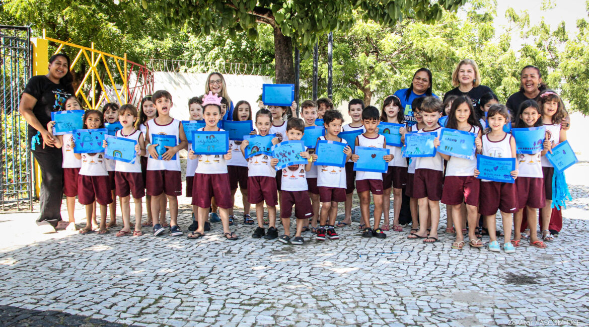 Várias crianças posando para foto, sorrindo e exibindo cartazes na cor azul. Atrás delas, cinco mulheres dando o apoio