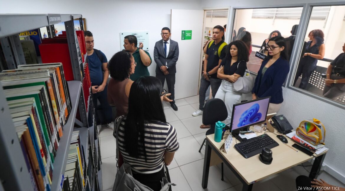Foto com mulheres e homens em uma sala pequena, em pé. Ao lado deles, uma estande com vários livros e uma mesa com um computador