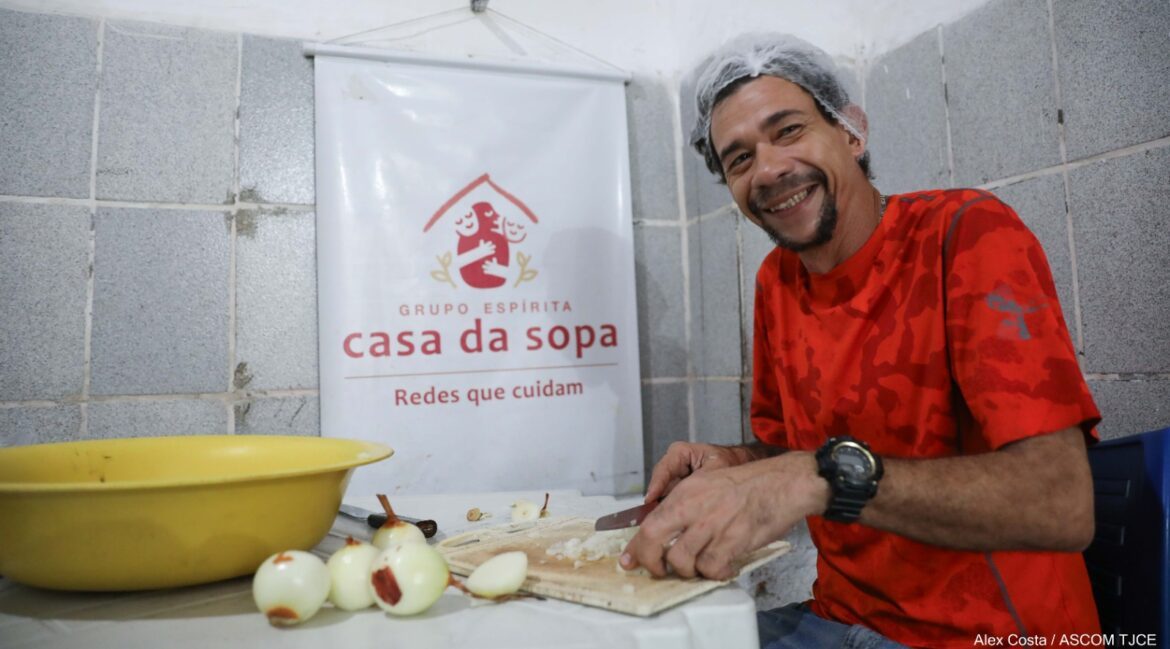 Foto de um homem sorrindo, sentado, enquanto corta cebolas em uma mesa