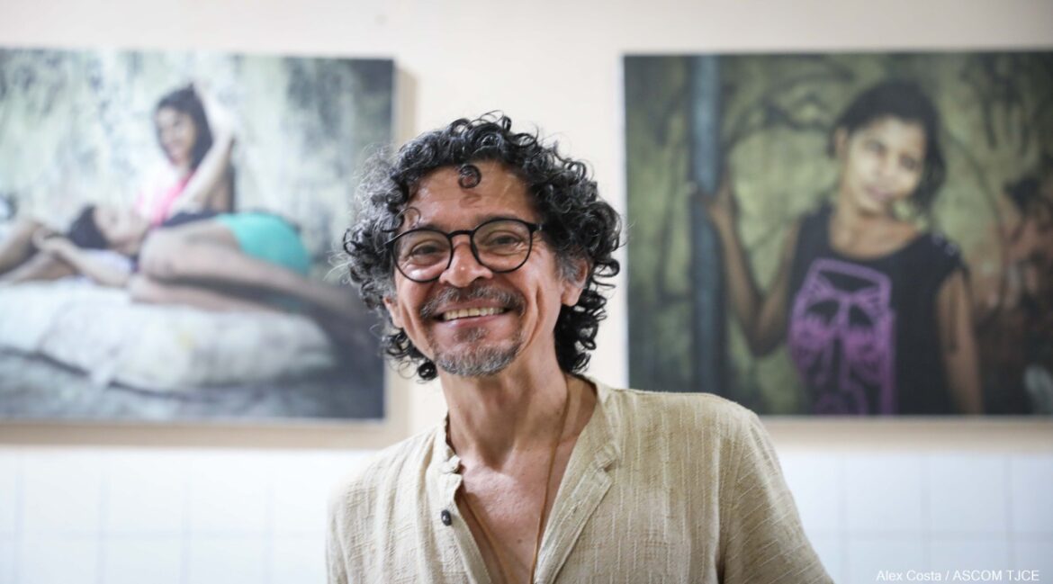 Foto de um homem sorrindo e, ao fundo, dois quadros na parede retratando pessoas 