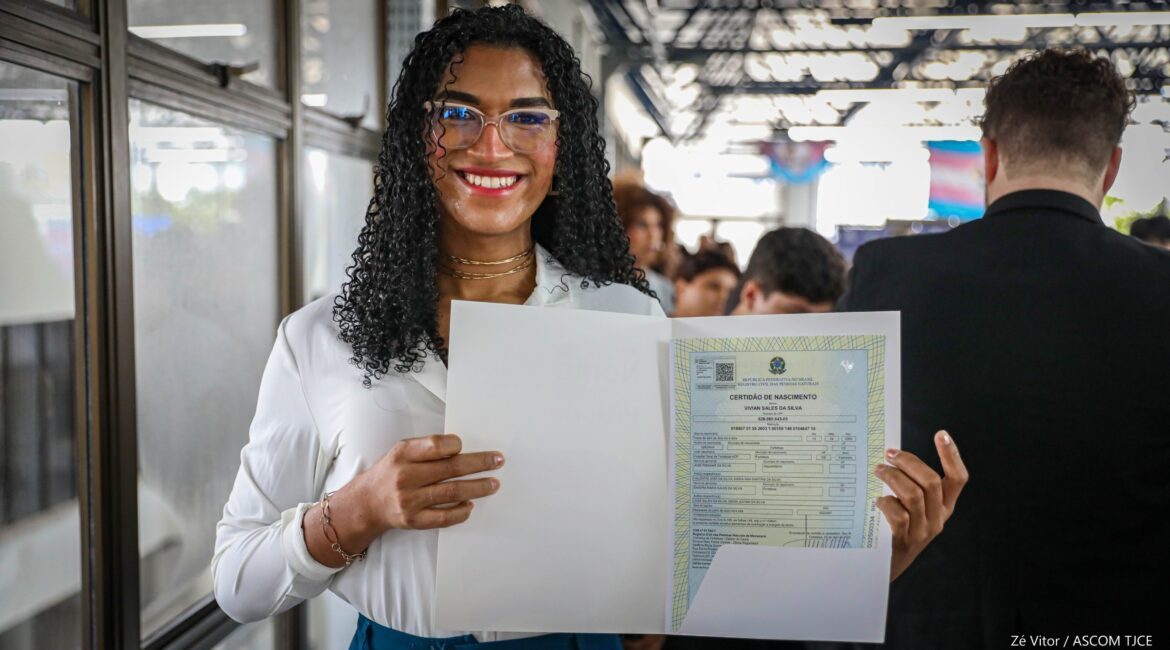 Mulher sorrindo, em pé, exibindo o documento de registro civil