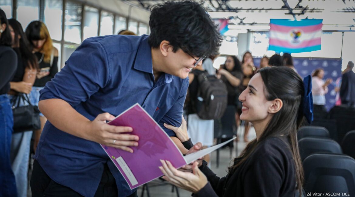 Uma pessoa sorrindo, em pé, exibindo o documento para uma outra que está sentada, sorrindo também