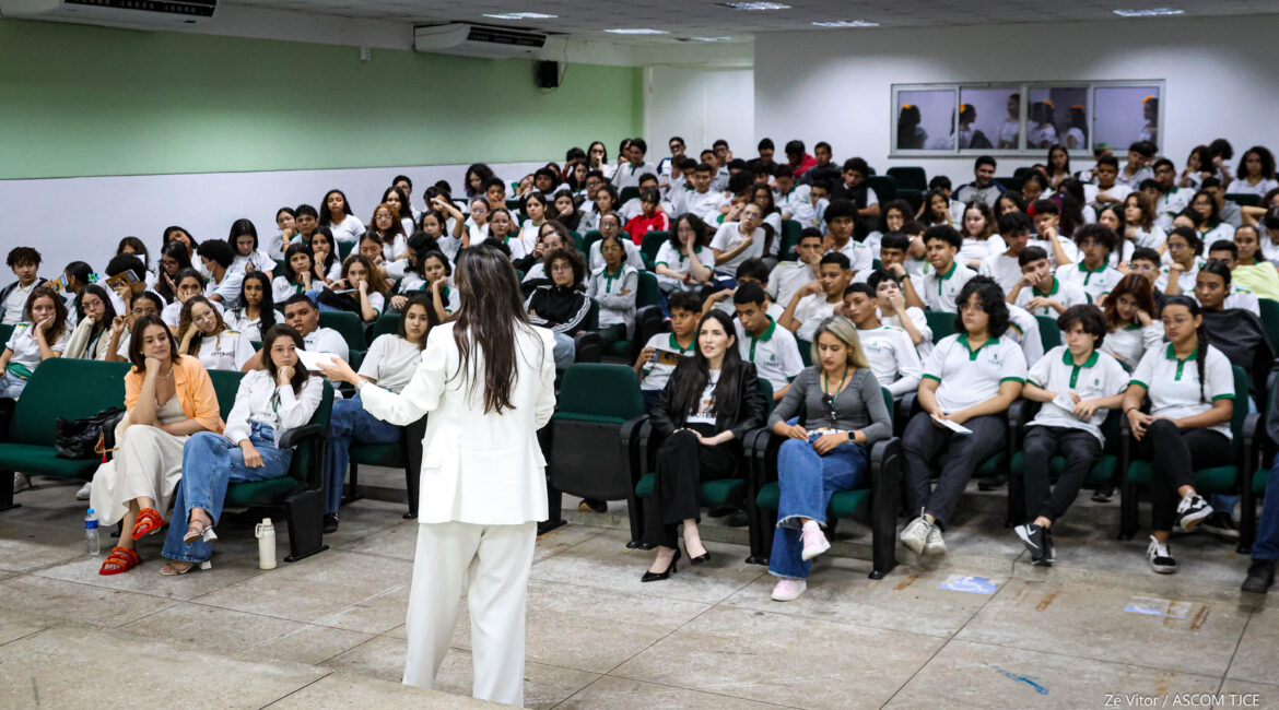 Foto de uma mulher de branco, de costas, falando ao microfone para vários alunos e alunas na plateia