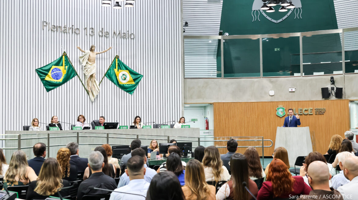 Várias pessoas sentadas em um auditório e, à frente delas, mais pessoas sentadas próximas a uma grande bancada. À direita delas, um homem falando ao microfone, no púlpito Várias pessoas sentadas em um auditório e, à frente delas, mais pessoas sentadas próximas a uma grande bancada. À direita delas, um homem falando ao microfone, no púlpito