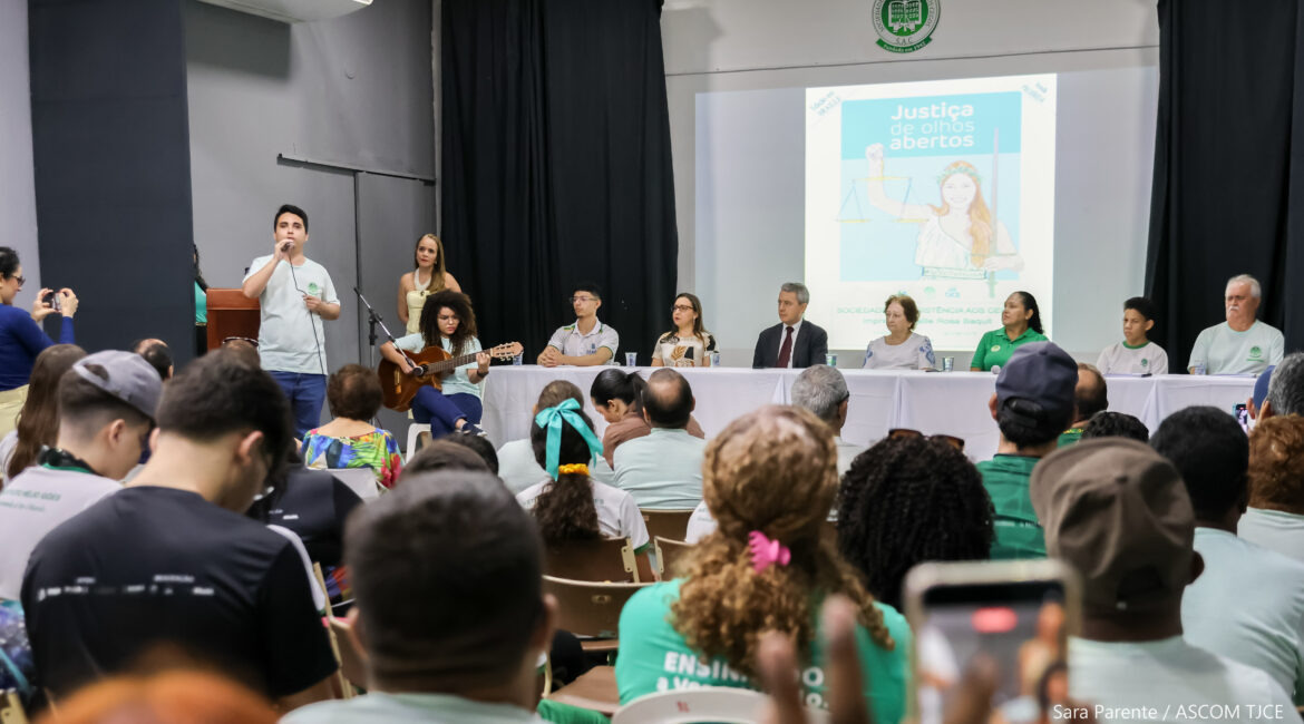 Foto em que aparecem parte do público sentado, a composição da mesa com sete pessoas sentadas, e dois jovens fazendo apresentação musical: um canta ao microfone e a outra toca violão