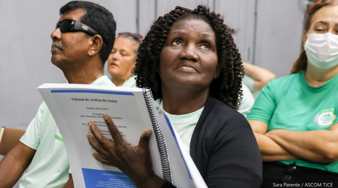 Uma mulher segurando um exemplar da cartilha em Braille, olhando para o alto. Ao dela, um homem de óculos escuros e, atrás dela, uma mulher usando máscara