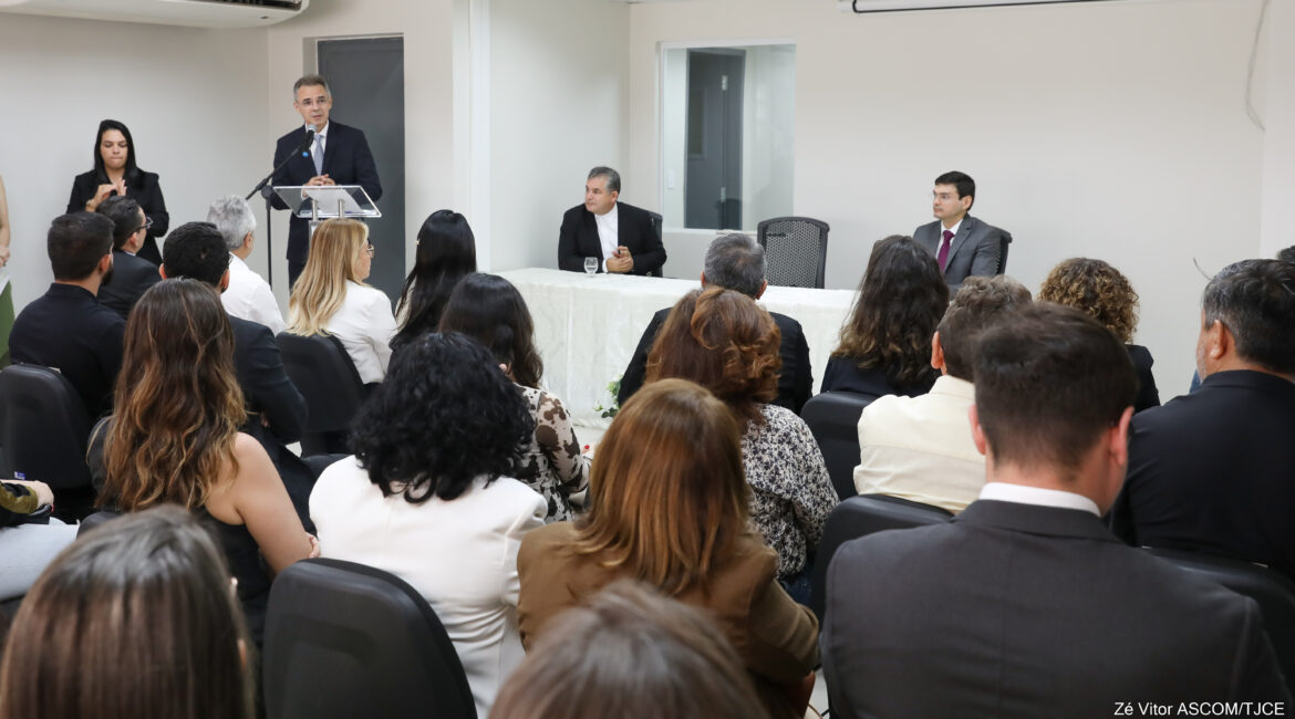 Homem falando ao microfone, em pé, numa sala. Na frente dele, várias pessoas sentadas ouvindo-o
