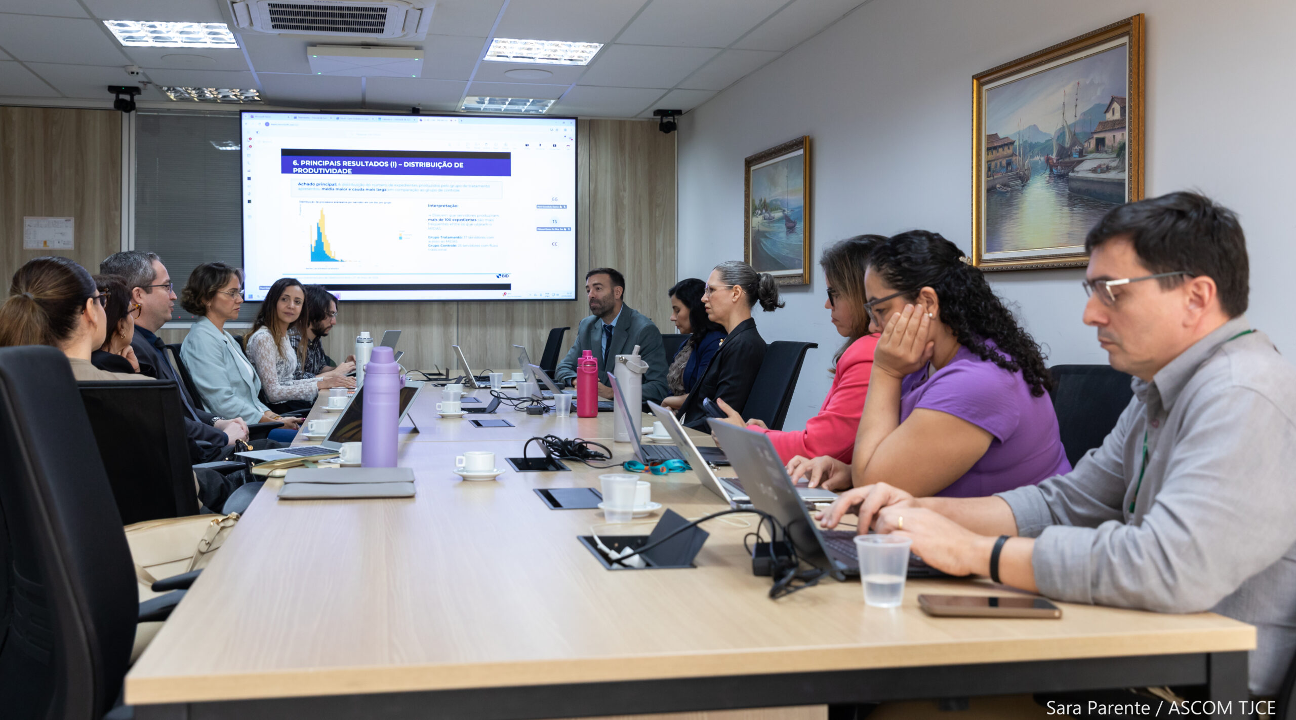 Pessoas em uma mesa comprida de reuniões, com telão com projeção ao fundo.