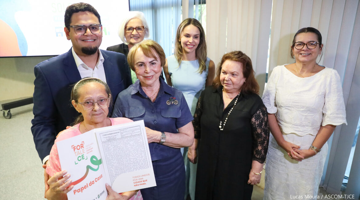 Foto com seis mulheres e um homem sorrindo para foto. Uma delas, mostra satisfeita um doumento 