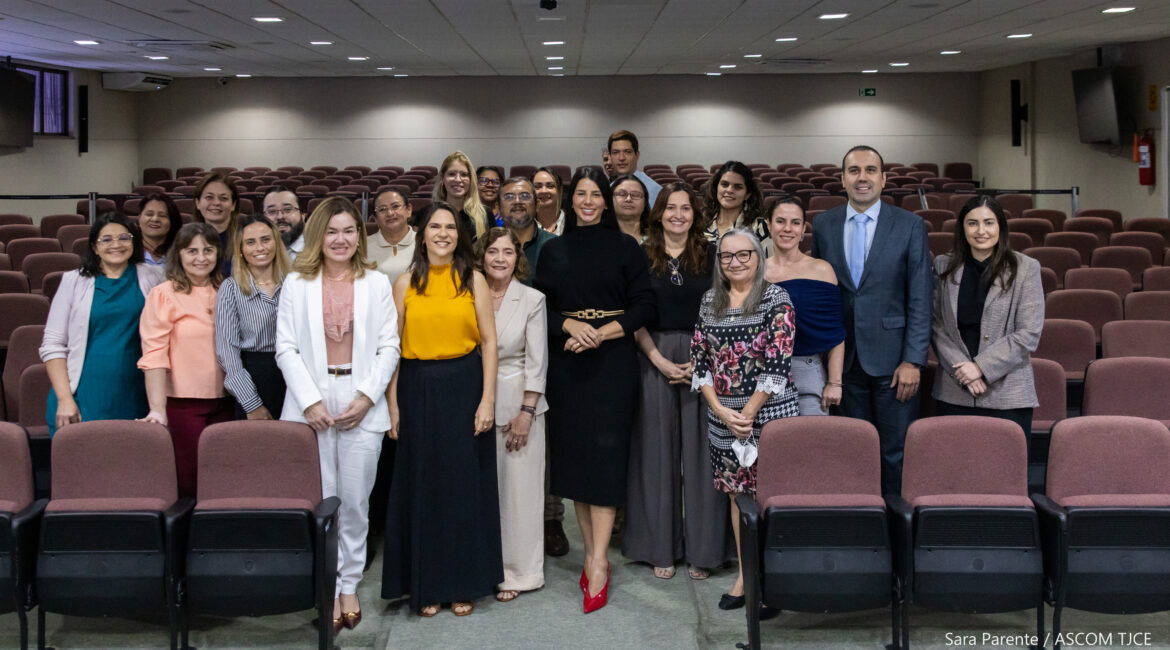 Imagem de várias pessoas posando para foto, em pé, sorrindo, em um auditório