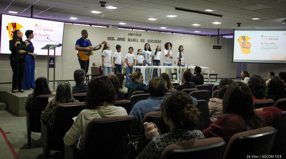 Várias crianças e adoldescentes perfilhadas no palco durante apresentação musical. Ao lado delas, um homem toca no violão