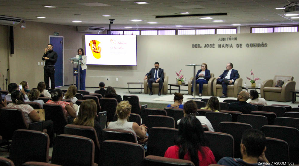 Imagem de um auditório em que se vê várias pessoas sentadas na plateia de costas e, no palco, dois homens e uma mulher sentada ao centro, de frente. Ao lado delas, uma mulher falando ao microfone, em pé e, ao lado desta, um homem realizando o trabalho de intérprete de Libras Imagem de um auditório em que se vê várias pessoas sentadas na plateia de costas e, no palco, dois homens e uma mulher sentada ao centro, de frente. Ao lado delas, uma mulher falando ao microfone, em pé e, ao lado desta, um homem realizando o trabalho de intérprete de Libras