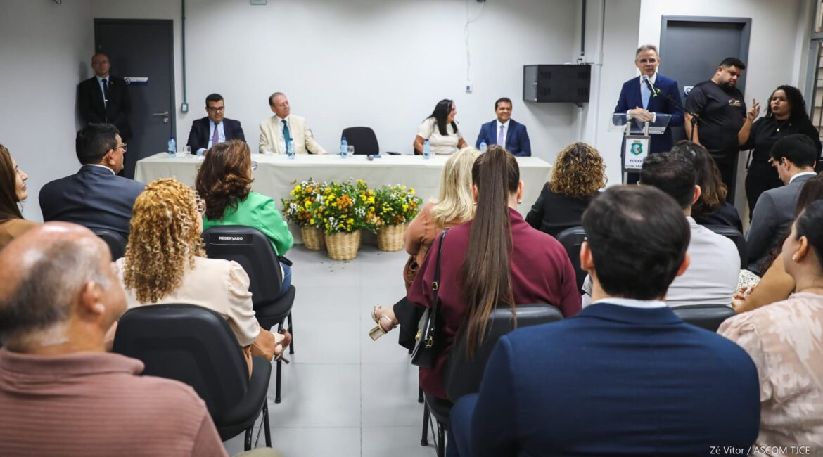 Homem falando ao microfone, em pé, no auditório. Ao lado dele, uma mesa em que estão sentados três homens e uma mulher. À frente dele, várias pessoas ouvindo sentadas Homem falando ao microfone, em pé, no auditório. Ao lado dele, uma mesa em que estão sentados três homens e uma mulher. À frente dele, várias pessoas ouvindo sentadas