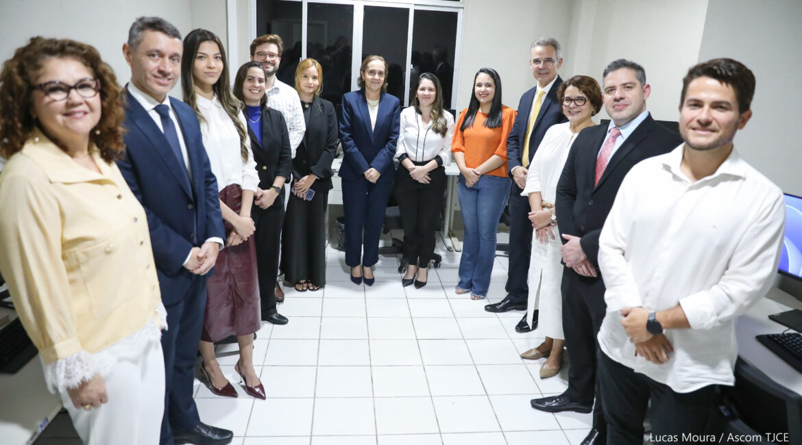 Imagem de várias pessoas posando para foto no formato de U, sorrindo, em pé, em uma sala