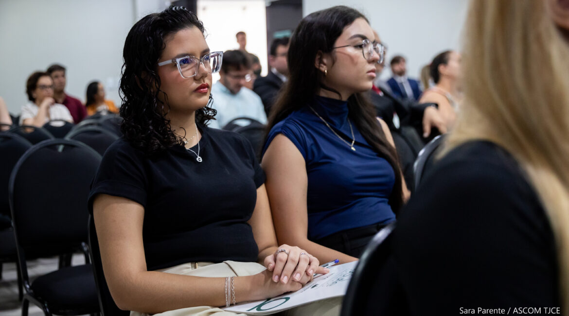 Foto que traz em destaque duas jovens, sentadas, na plateia, ouvindo as explicações Foto que traz em destaque duas jovens, sentadas, na plateia, ouvindo as explicações