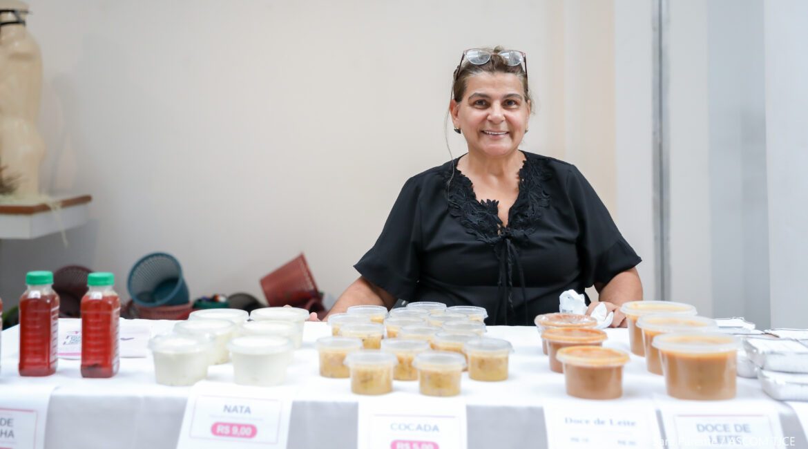 Mulher sorrindo, sentada, vendendo produtos comestíveis que estão em cima da mesa, produzidos por ela Mulher sorrindo, sentada, vendendo produtos comestíveis que estão em cima da mesa, produzidos por ela