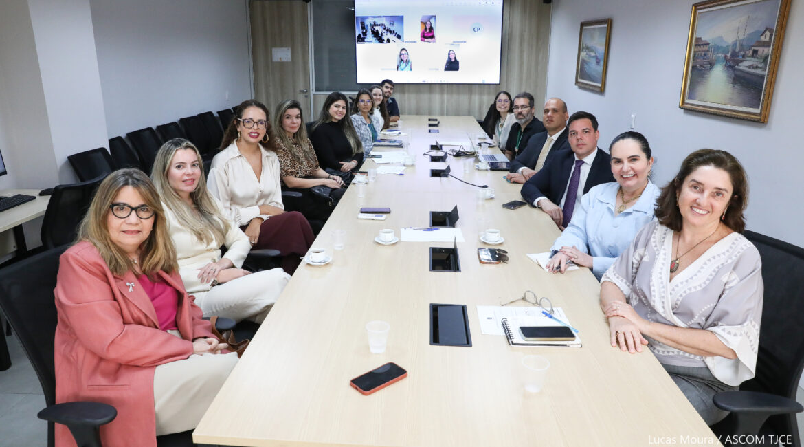 Imagem de mulheres e homens sorrindo, posando para foto, sentados, ao redor de uma grande mesa de trabalho
