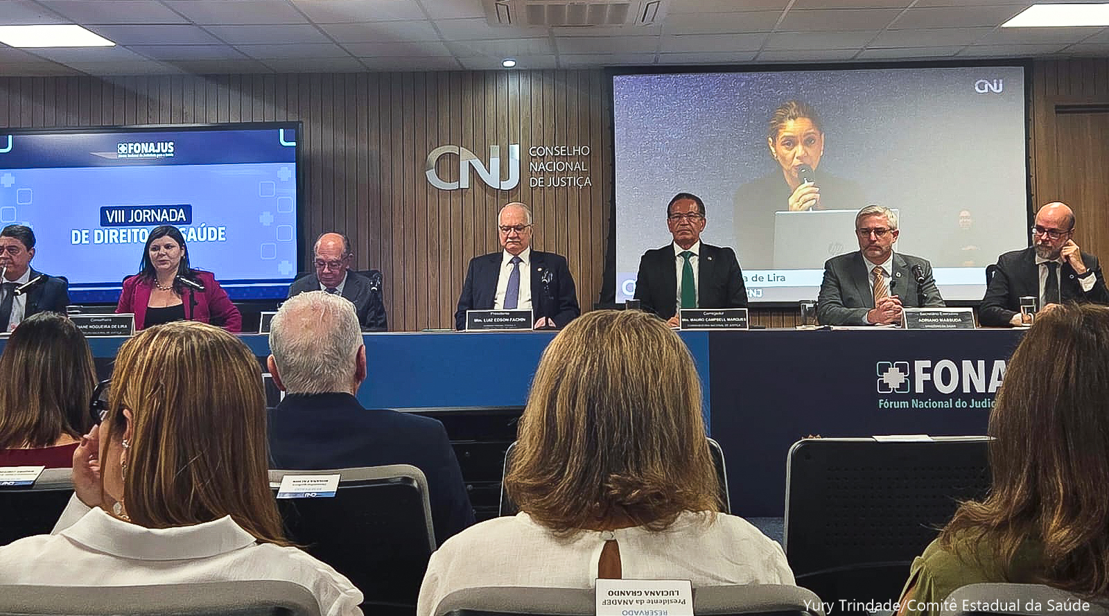 Mesa de trabalho com várias pessoas sentadas. À frente delas, pessoas sentadas em um grande auditório