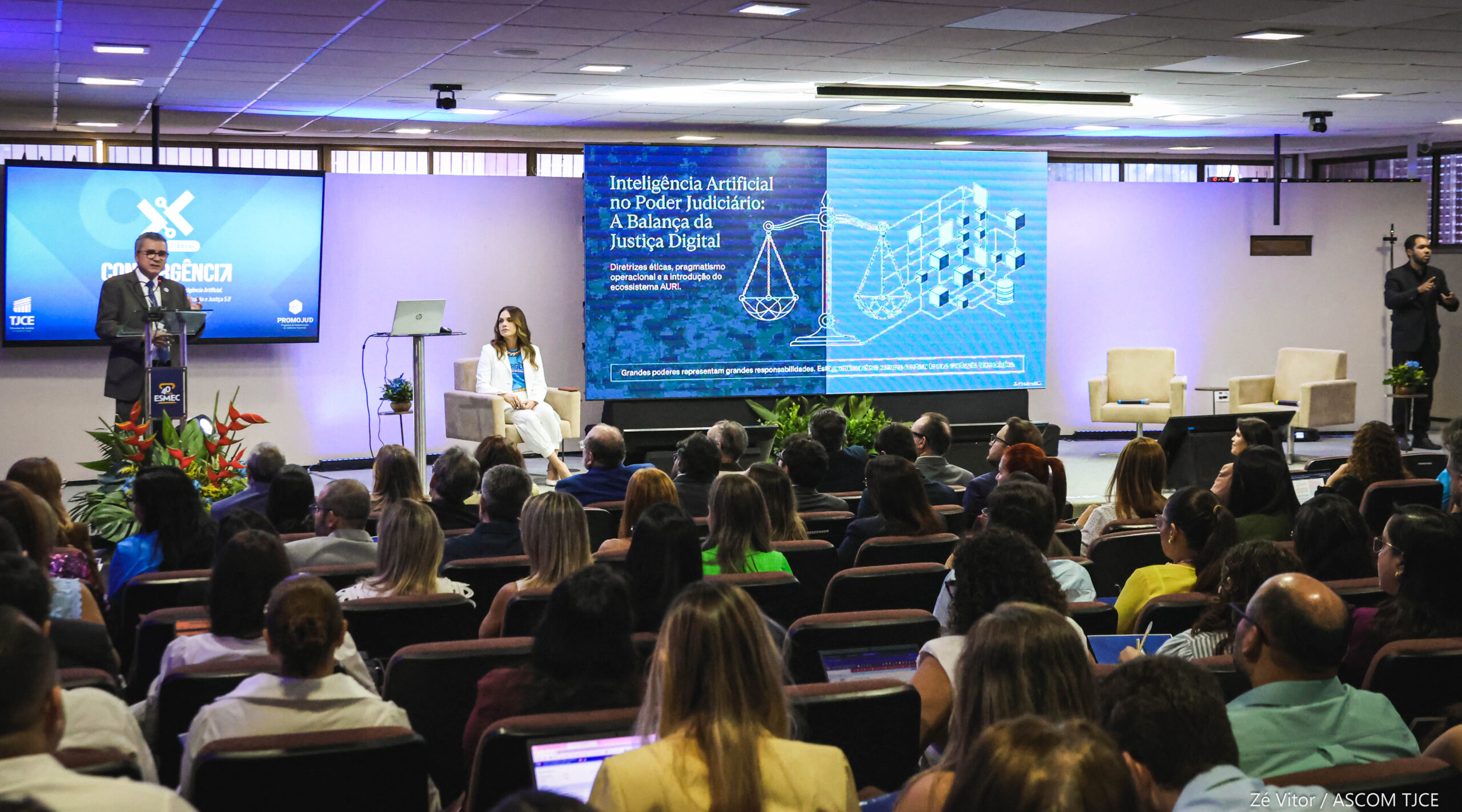 Foto de um homem falando ao microfone no palco e, ao lado dele, uma mulher observando-o. Na frente deles, várias pessoas na plateia sentadas